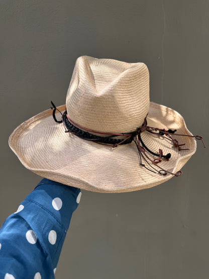 Beige straw hat with decorative band held by a person wearing a blue polka dot shirt against a gray background